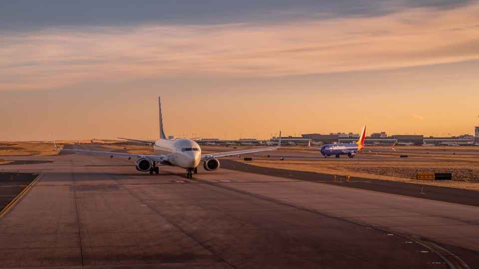 Twee passagiersvliegtuigen taxiën over de landingsbaan van een luchthaven tijdens een oranjegekleurde zonsondergang.