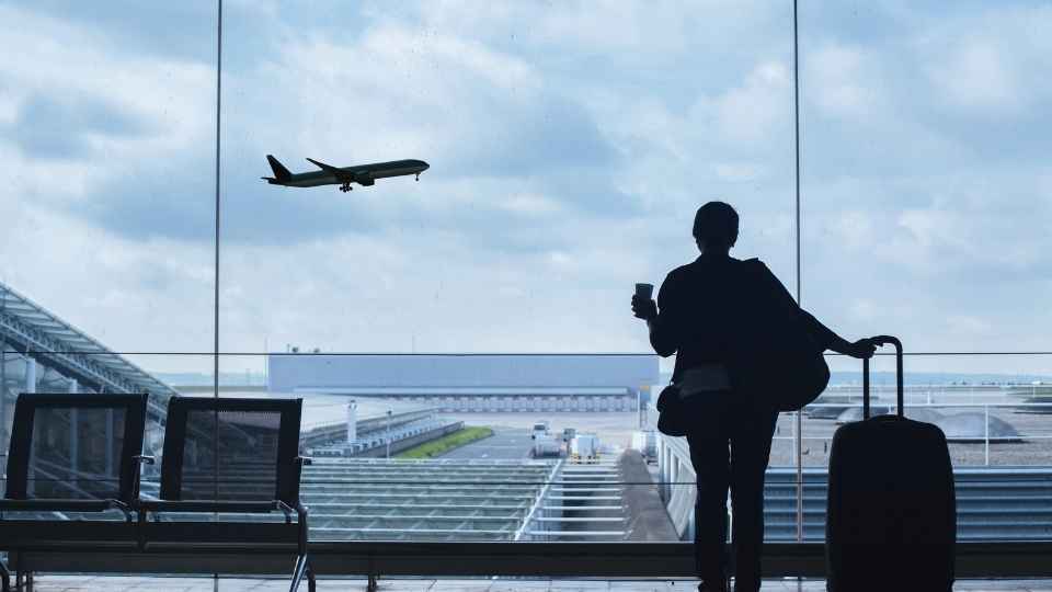 Reiziger met koffiebeker en koffer kijkt door het raam van de luchthaven naar een opstijgend vliegtuig.
