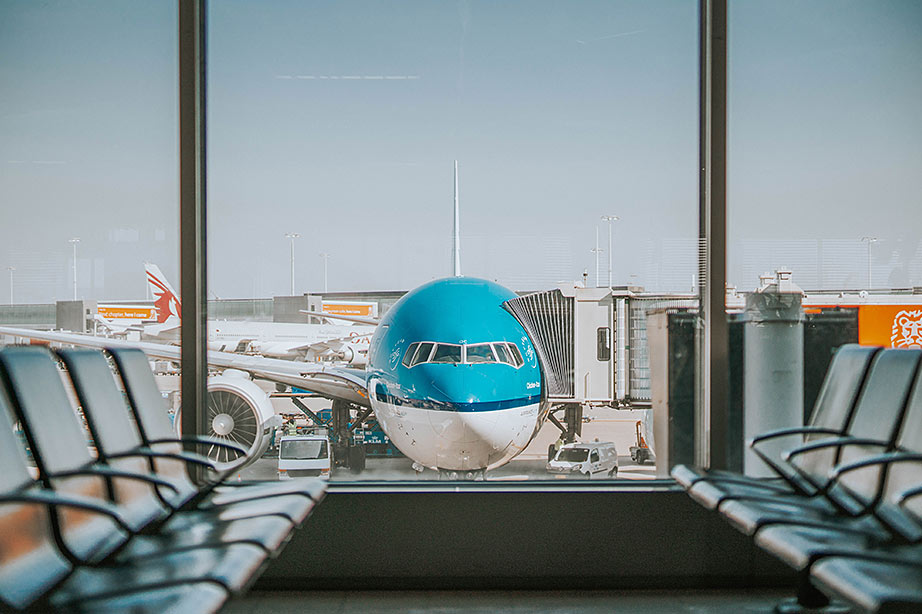 Een KLM-vliegtuig staat geparkeerd aan de gate van een luchthaven, gezien vanuit een lege wachtruimte met metalen stoelen.