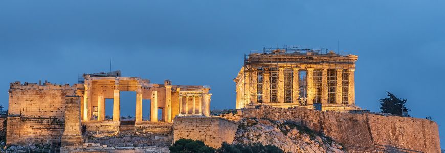 De verlichte ruïnes van de Akropolis in Athene, waaronder het Parthenon, tegen een blauwe schemerlucht.