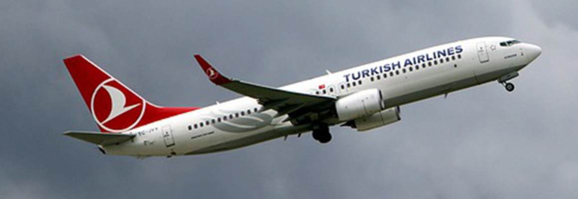 Een vliegtuig van Turkish Airlines, met een wit lichaam en het rode logo van de maatschappij op de staart, is zichtbaar in de lucht tijdens een heldere dag met enkele wolken op de achtergrond.