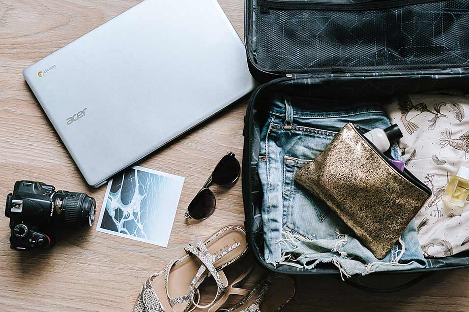 Open koffer met zomerkleding, sandalen, zonnebril, toilettas en reisbenodigdheden zoals een laptop en camera op een houten vloer, klaar voor vertrek.