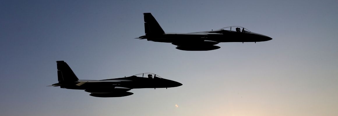 Twee gevechtsvliegtuigen vliegen in formatie tijdens zonsondergang, silhouetten duidelijk zichtbaar tegen een heldere hemel.