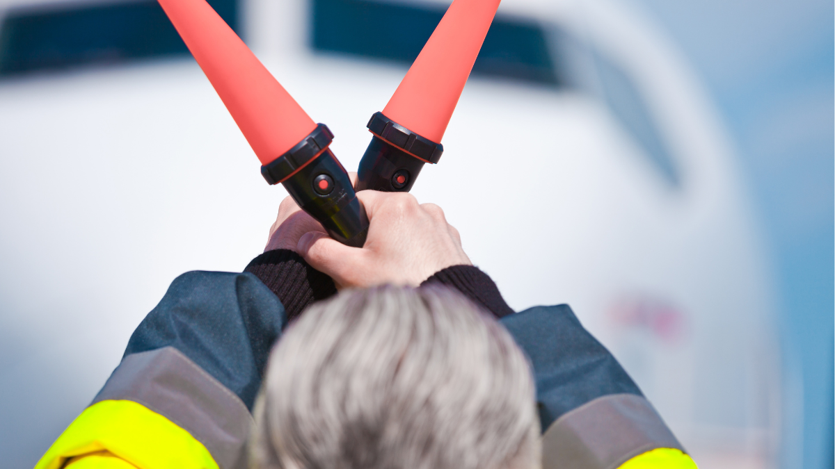 Grondmedewerker op een luchthaven houdt twee oranje verlichte marshallingsstaven gekruist omhoog, met een vliegtuig op de achtergrond.