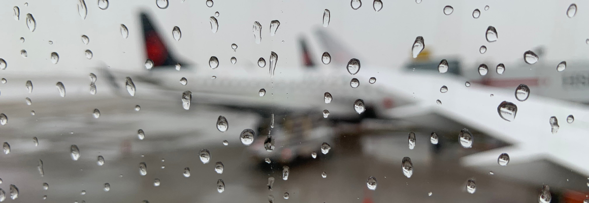 Uitzicht vanuit een vliegtuigraam op een natte luchthaven, met regendruppels op het raam en een onscherp vliegtuig op de achtergrond.