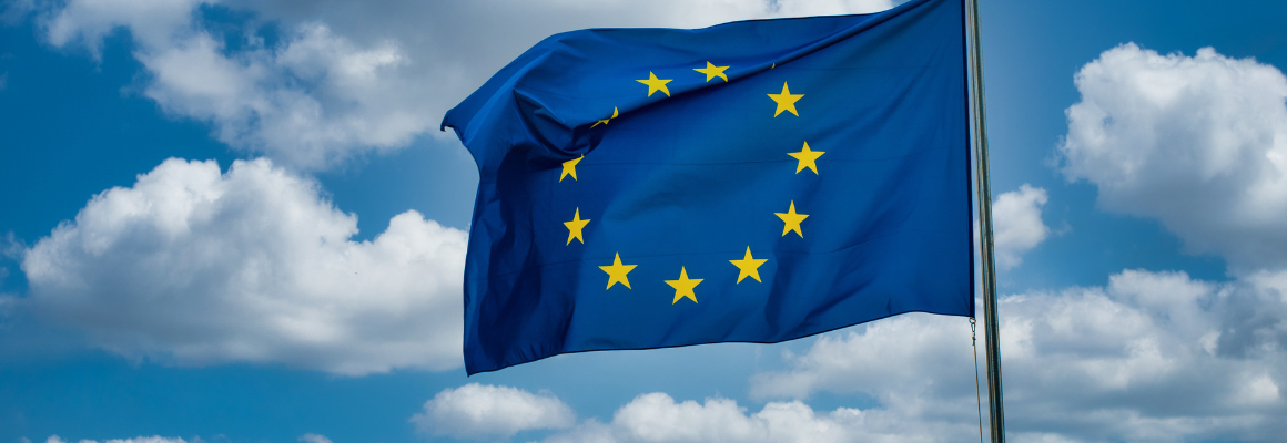Blauwe vlag met twaalf gele sterren in een cirkel, wapperend in de wind tegen een blauwe lucht met witte wolken (vlag van de Europese Unie).